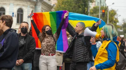 One person holding up a LGBTQ Pride flag next to a person holding up a Ukrainian Flag and someone speaking into a megaphone