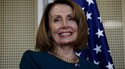 Speaker of the United States House of Representatives Nancy Pelosi smiles while wearing a blue suit and standing in front of an American flag in Brussels, Belgium