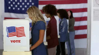 Four women of various demographics filling in ballots and casting votes in booths at polling station, US flag on wall at back.