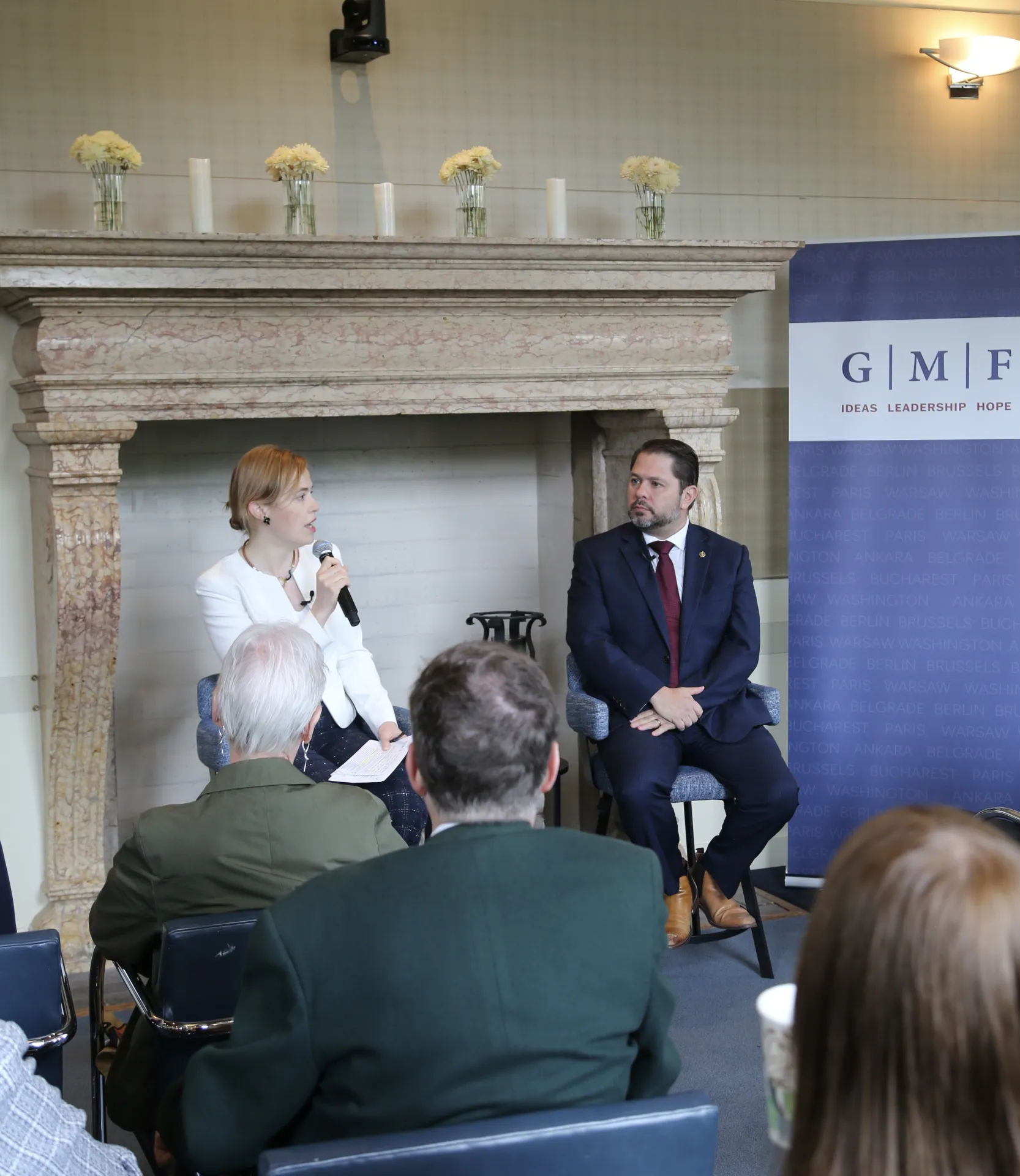 A man and a woman speaking at the front of a room in front of a fireplace