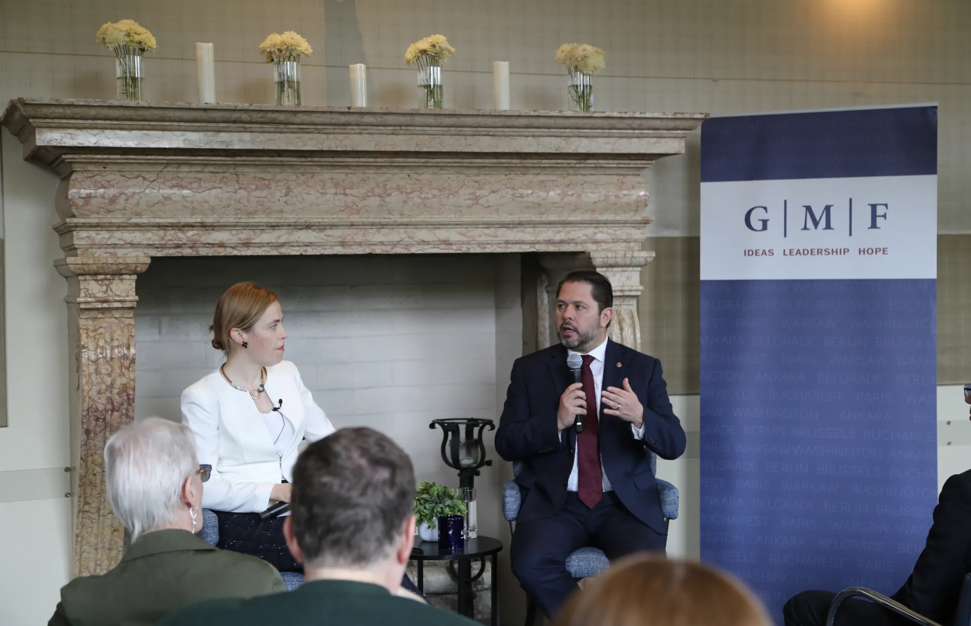A man and a woman speaking at the front of a room in front of a fireplace