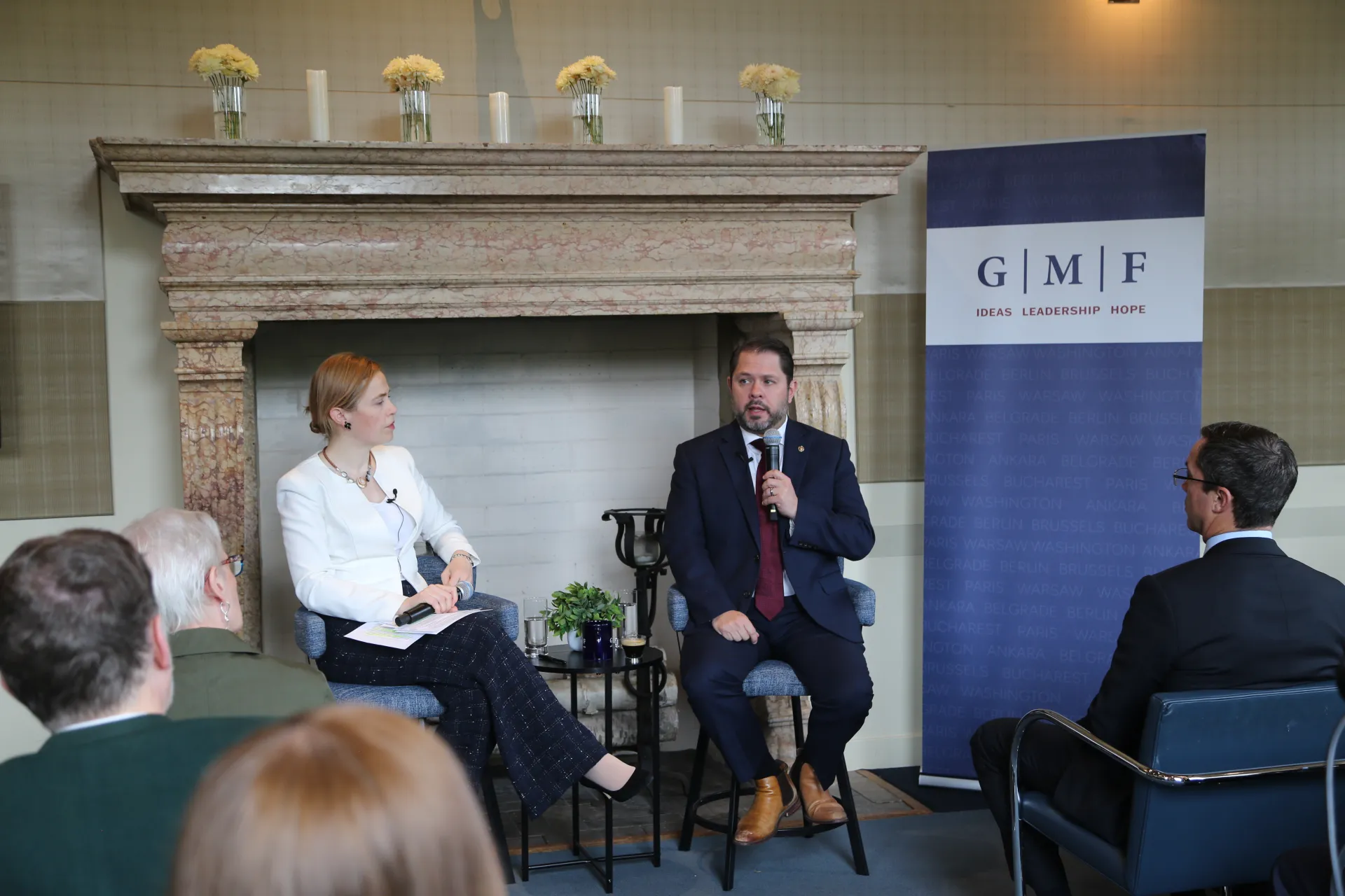 A man and a woman speaking at the front of a room in front of a fireplace