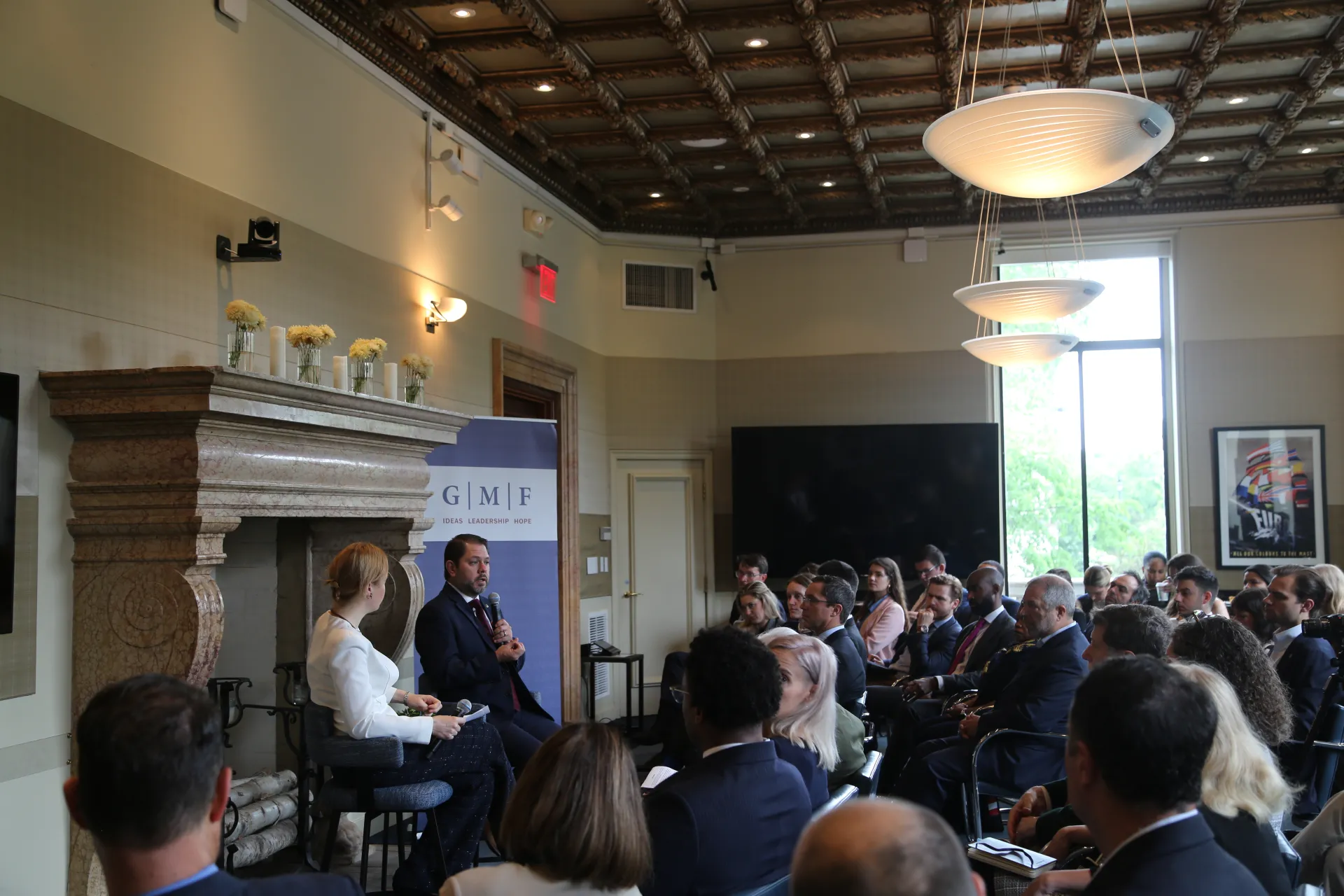 A man and a woman speaking at the front of a room in front of a fireplace